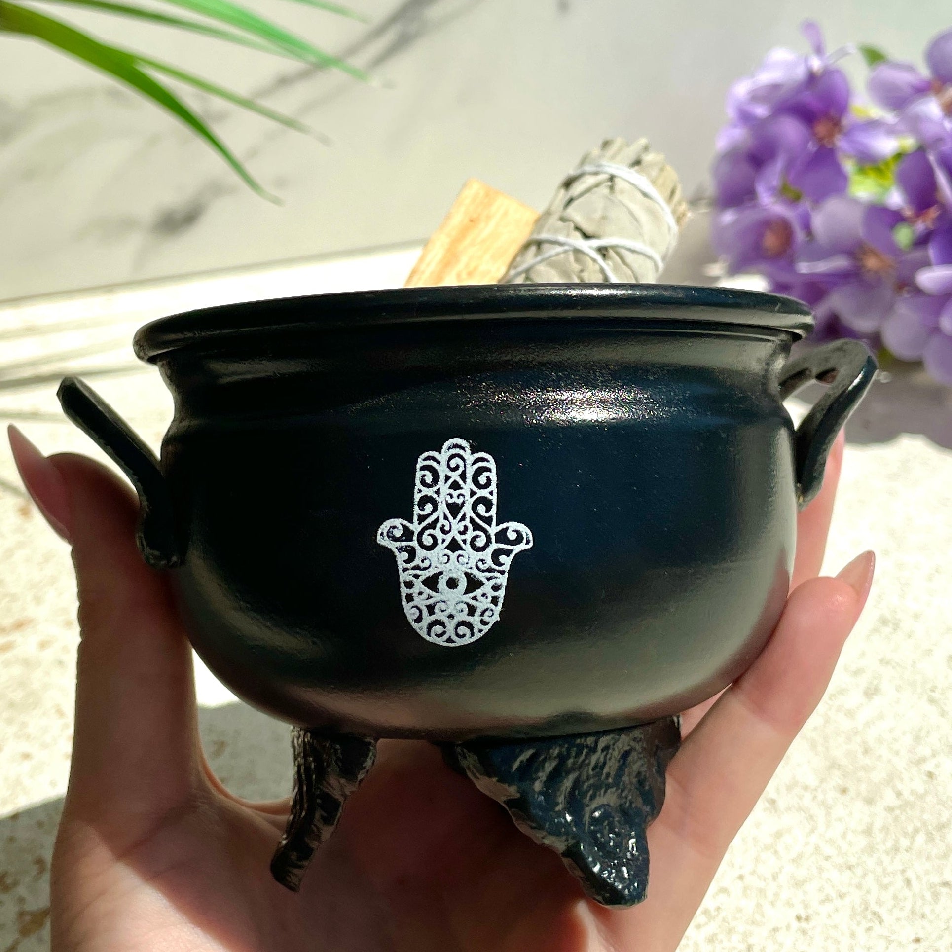 Black cauldron with Hamsa hand design held in a hand, with plants and flowers in the background.